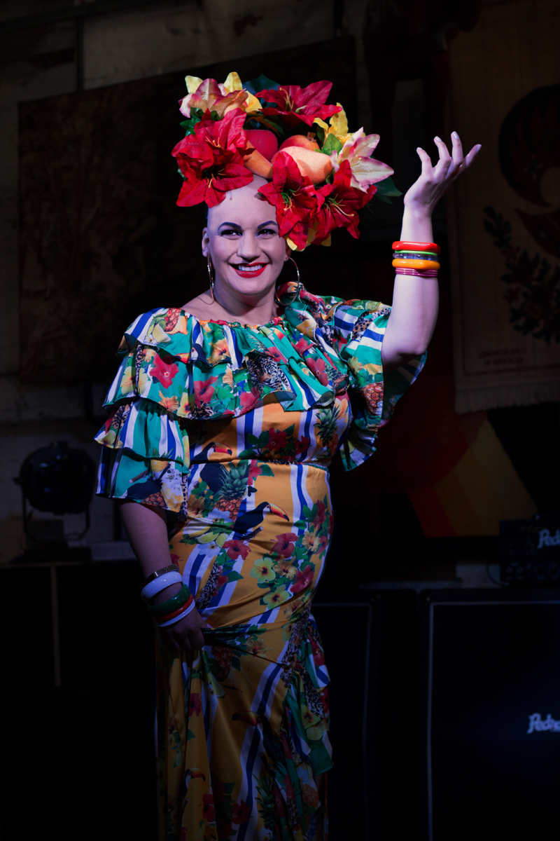Mulher branca de cabelo raspado segura levante o braço direito em um passo de dança que remete à Carmen Miranda.  Usa um vestido colorido, com temas florais, e um arranjo artificial de flores e frutas na cabeça.