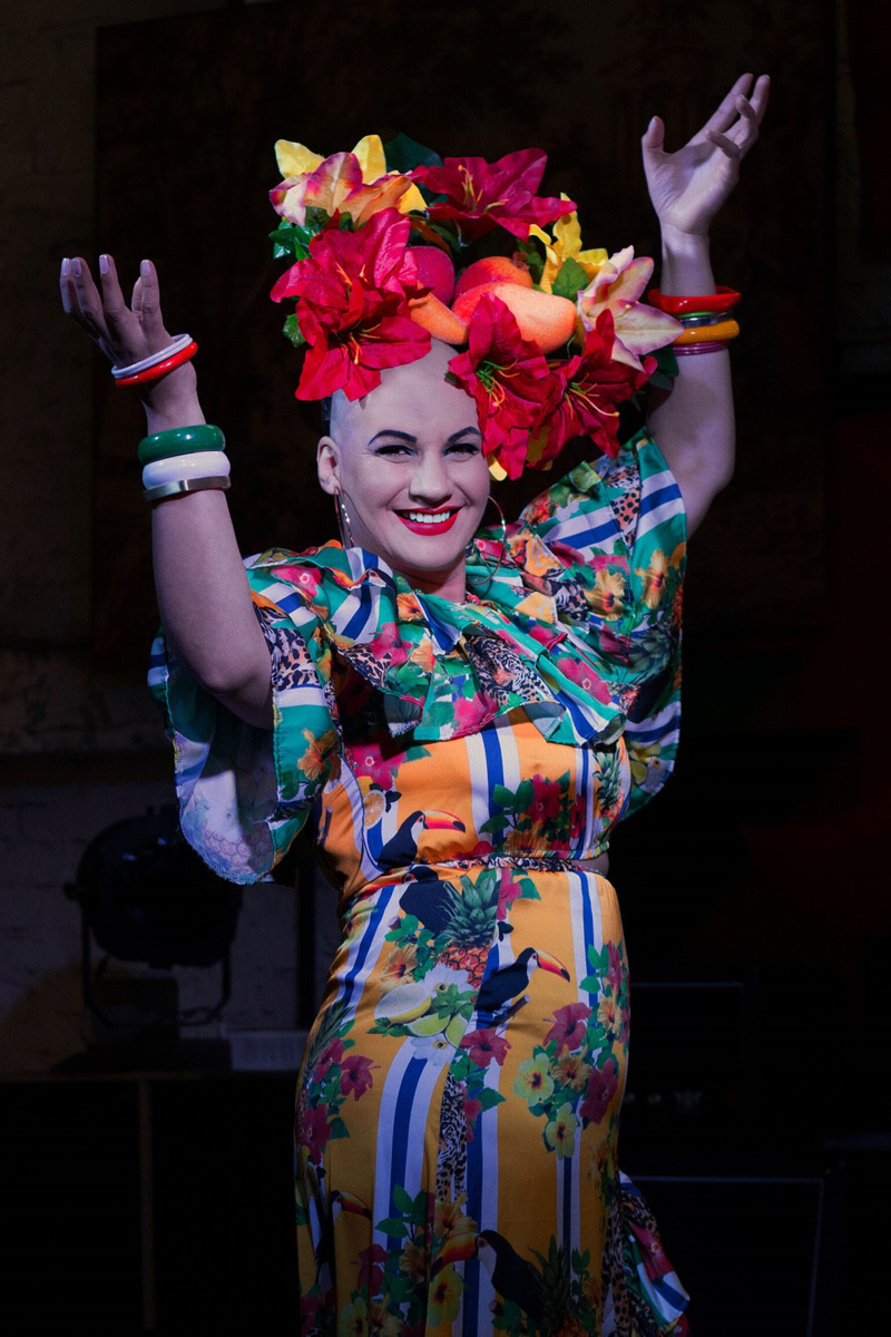 Mulher branca de cabelo raspado segura levanta os dois braços e sorri.  Usa um vestido colorido, com temas florais, e um arranjo artificial de flores e frutas na cabeça. O estilo remete à Carmen Miranda.