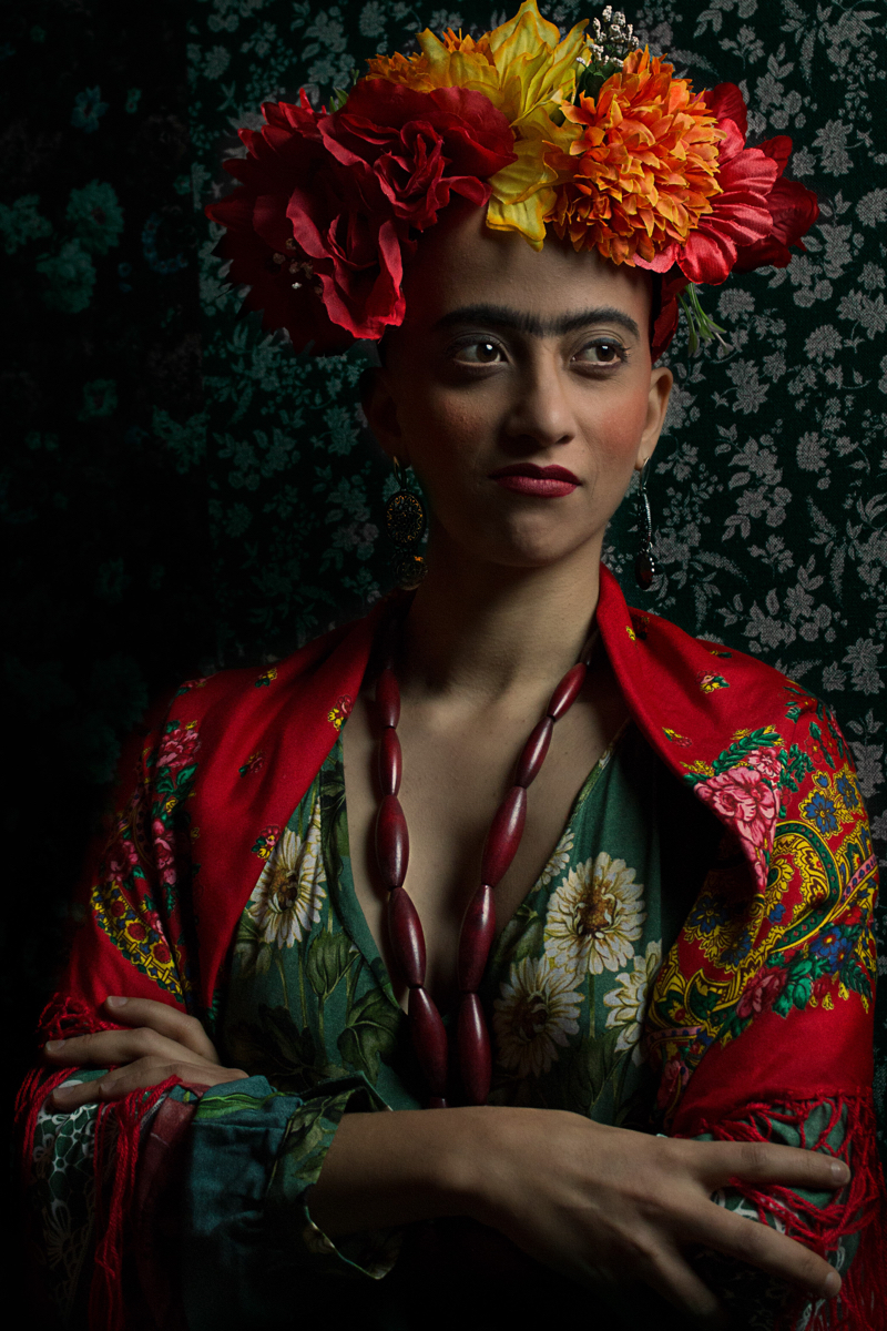 Mulher parda de cabelo raspado, sobrancelhas espessas e escuras. Usa flores vermelhas, laranjas e amarelas na cabeça e um vestido floral. Remete à pintora Frida Kahlo.