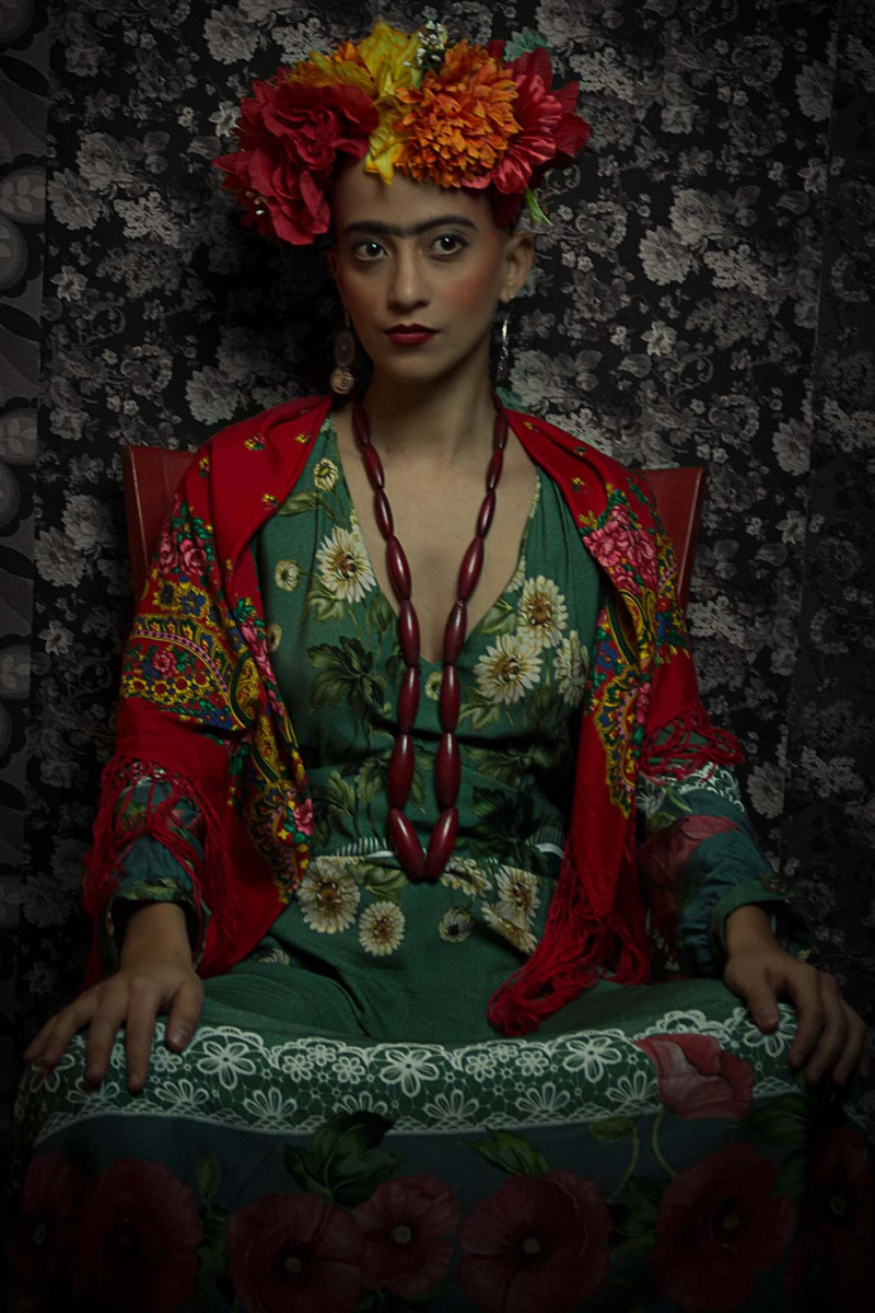 Mulher parda de cabelo raspado, sobrancelhas espessas e escuras. Usa flores vermelhas, laranjas e amarelas na cabeça e um vestido floral. Remete à pintora Frida Kahlo.
