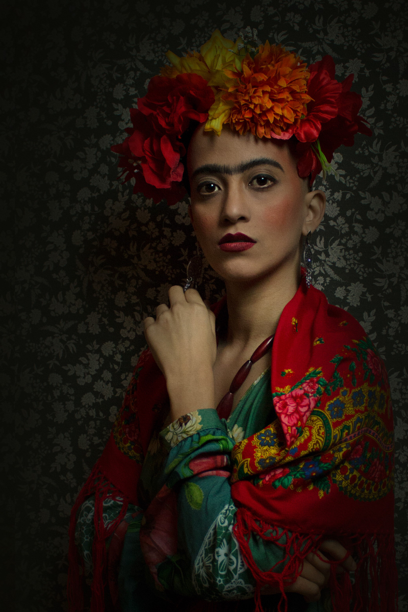 Mulher parda de cabelo raspado, sobrancelhas espessas e escuras. Usa flores vermelhas, laranjas e amarelas na cabeça e um vestido floral. Remete à pintora Frida Kahlo.
