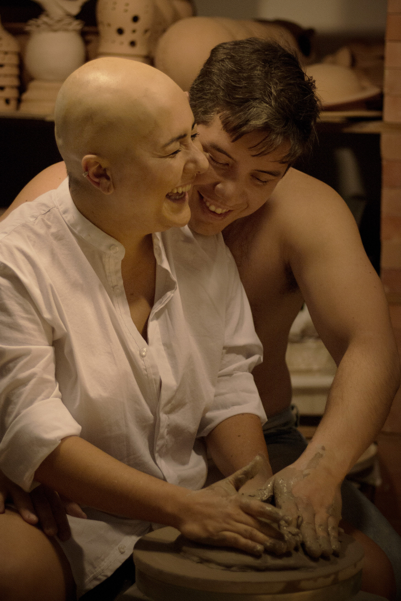 Mulher branca de cabelo raspada veste um camisa social branca e usa as mão para fazer um objeto de cerâmica ao lado de um homem sem camisa. A cena remete ao filme Ghost.