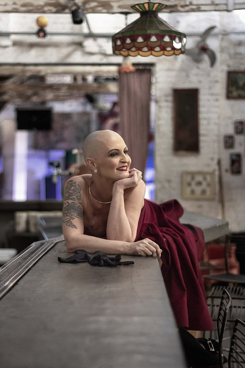 Mulher branca de cabelo raspada usa um vestido vermelho, batom vermelho, repousa os cotovelos sobre um balcão e olha para o lado sorrindo. Cena remeta à personagem Gilda.