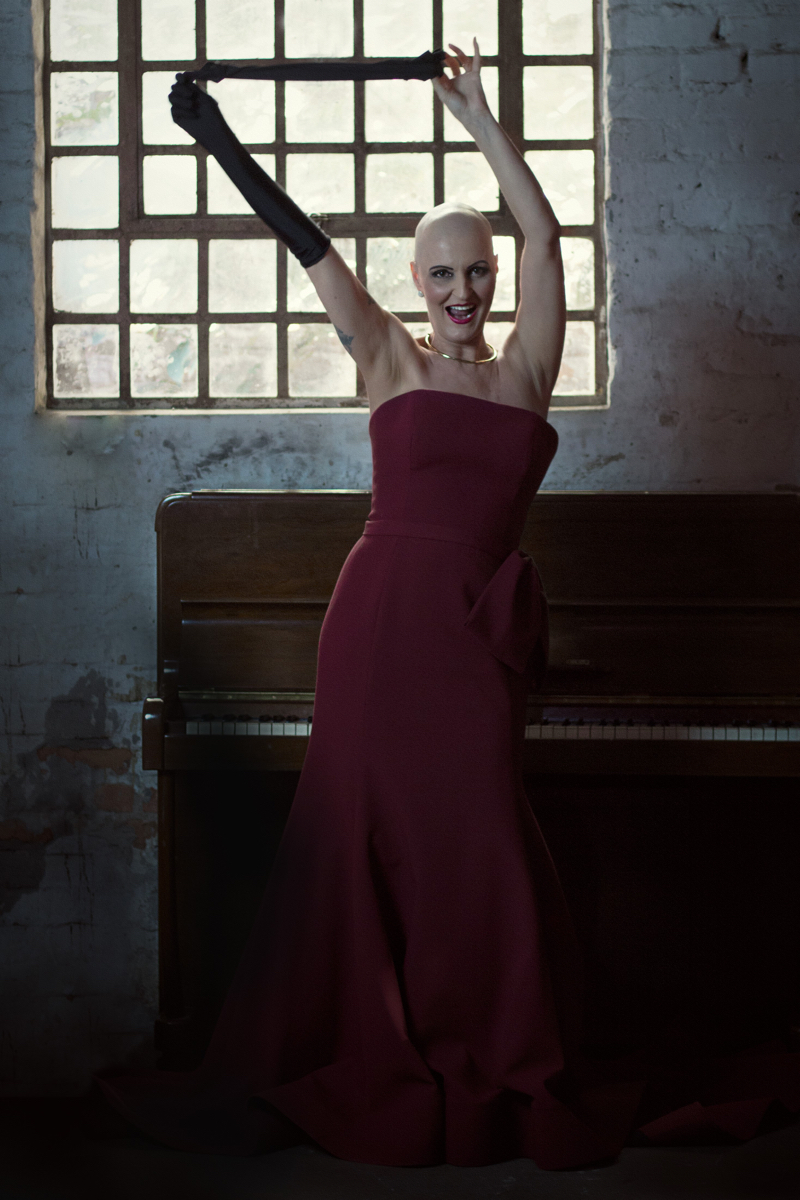 Mulher branca de cabelo raspada usa um vestido vermelho e batom vermelho. Está com os braços levantados. Usa uma luva preta comprida em uma mão, e retira a luva da outra. Cena remete à personagem Gilda.