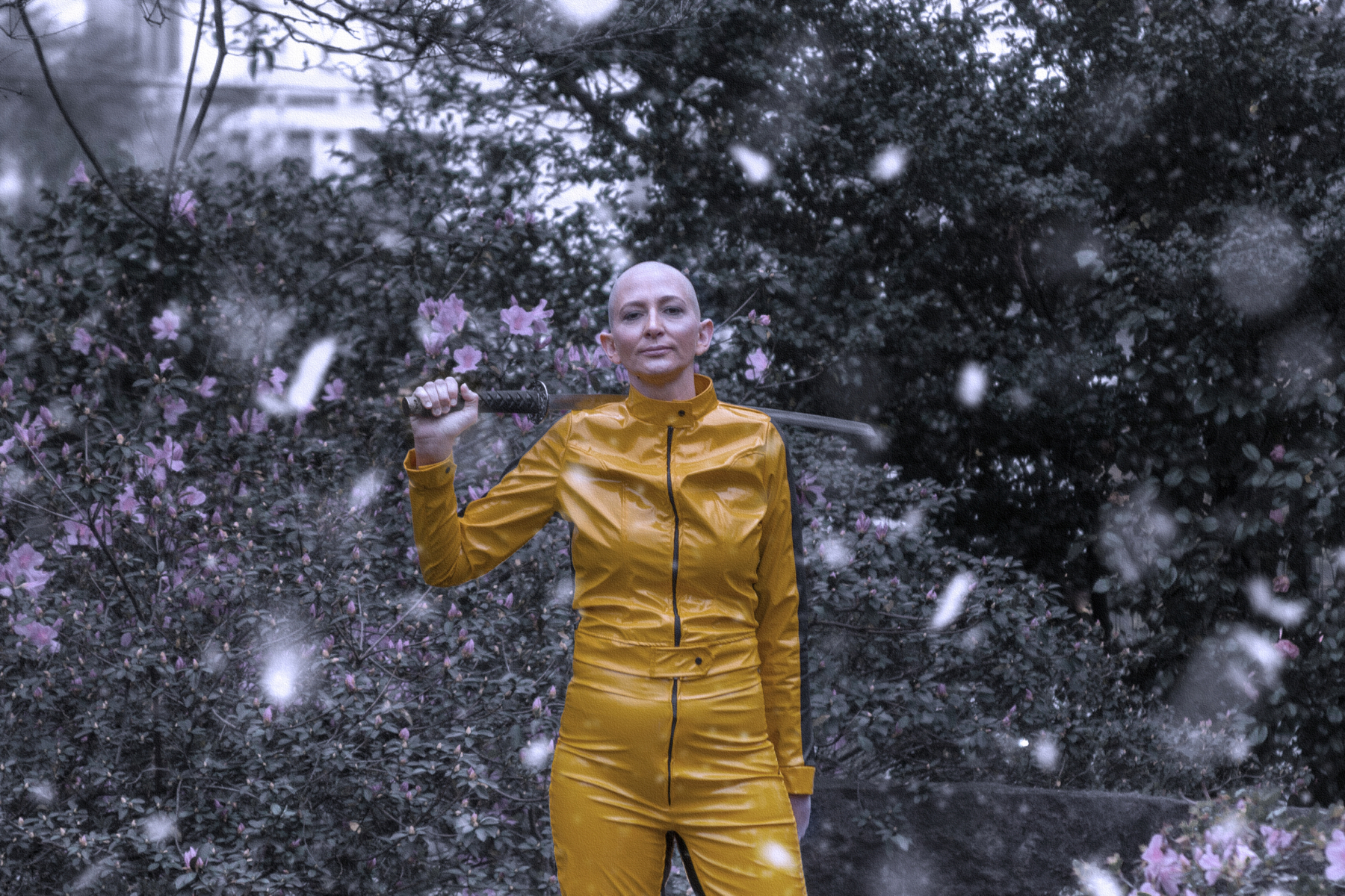 Mulher branca de cabelo raspada usa jaqueta e calça de couro amarelas. Segura uma katana que passa atrás dos seus ombros e olha para a câmera. Ao fundo há um arbusto com flores. Cena remete ao filme Kill Bill.