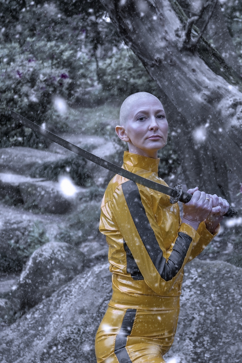 Mulher branca de cabelo raspada usa jaqueta e calça de couro amarelas. Está de lado e segura uma katana com as duas mãos, olhando para a câmera. A imagem simula neve, e há pedras e árvores ao fundo. Cena remeta ao filme Kill Bill.