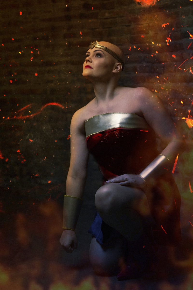 Mulher branca de cabelo raspado usa uma tiara dourada um uniforme de heróina em dourado, vermelho e azul. Esta de lado, apoiada em uma perna dobrada e olha para cima. Remete à Mulher Maravilha.