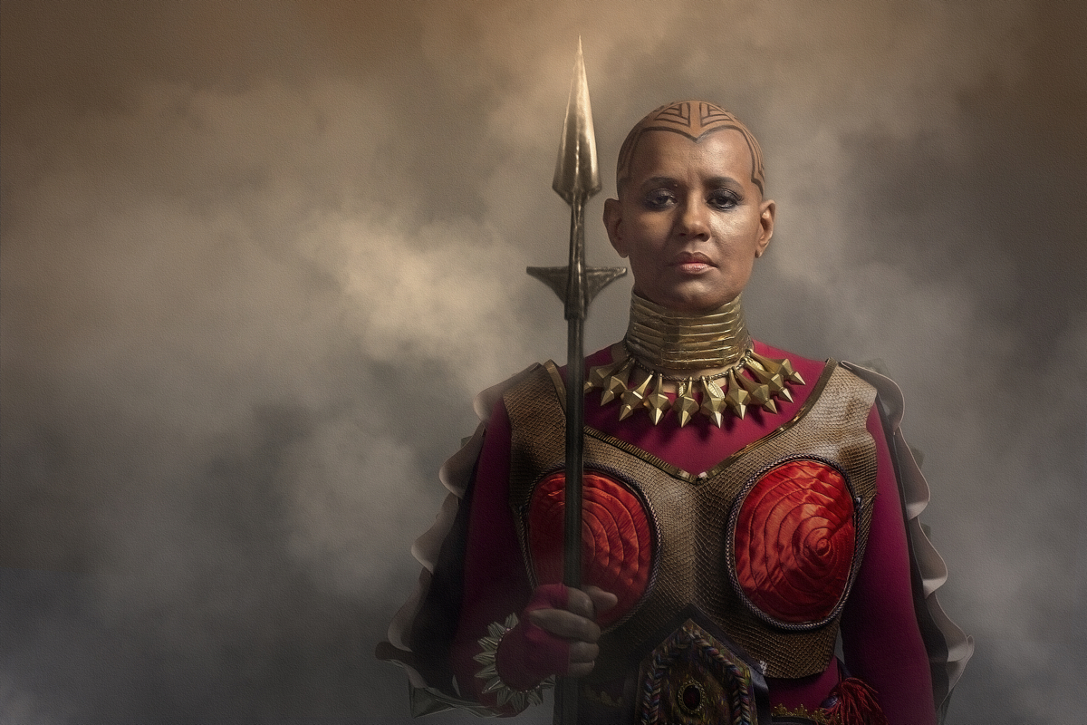 Retrato de uma mulher negra de cabelo raspado, empunhando uma lança dourada. Veste-se como uma guerreira, com colares dourados no pescoço e um uniforme vermelho e dourado. Remete à personagem Okoye.