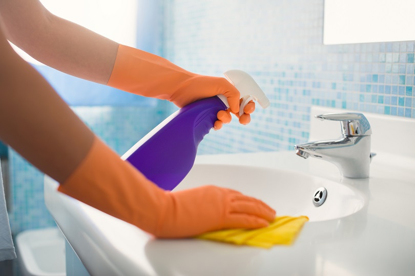 Foto ilustrativa das mãos de uma mulher limpando uma pia de banheiro. Ela está utilizando luvas e segura um produto de limpeza na mão direita. Com a mão esquerda, passa um pano de limpeza na superfície da pia.