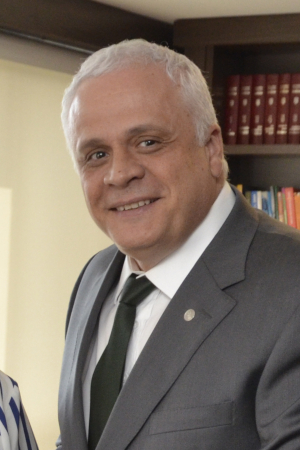 Retrato de Breier, sorrindo para a câmera. Ele tem pele branca, curtos cabelos brancos, olhos escuros, sem barba. Usa terno cinza, grava preta e camisa branca.