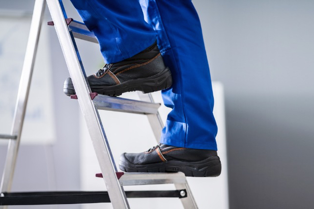 Foto ilustrativa dos pés de um homem subindo uma escada móvel. O homem está vestindo calça jeans e usando calçados.