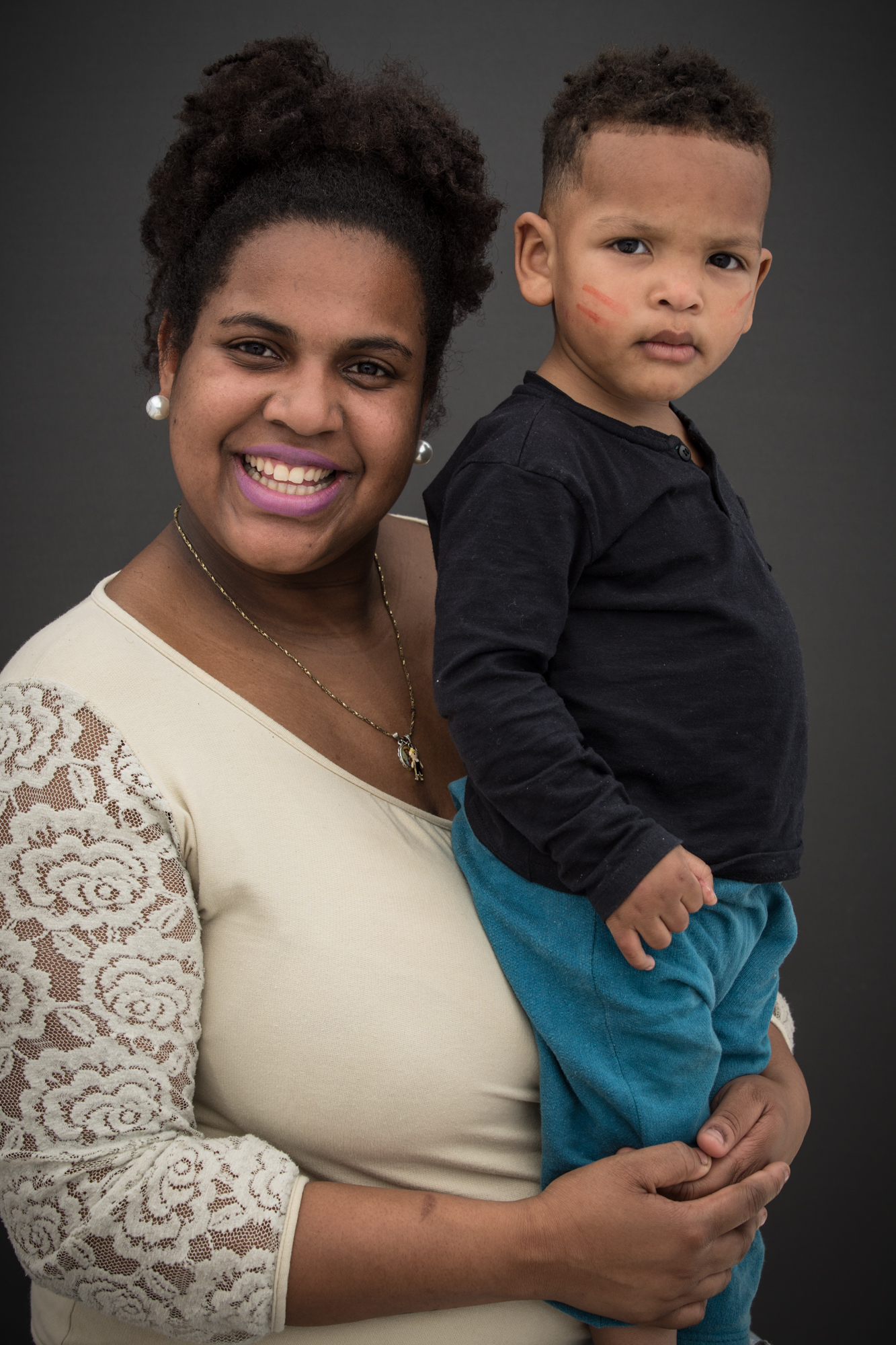Mulher negra segura seu filho. Ela tem o cabelo crespo com volume, usa brincos de pérola, colar dourado, batom e blusa bege. O menino está de pé, usa uma calça azul e camisa preta.
