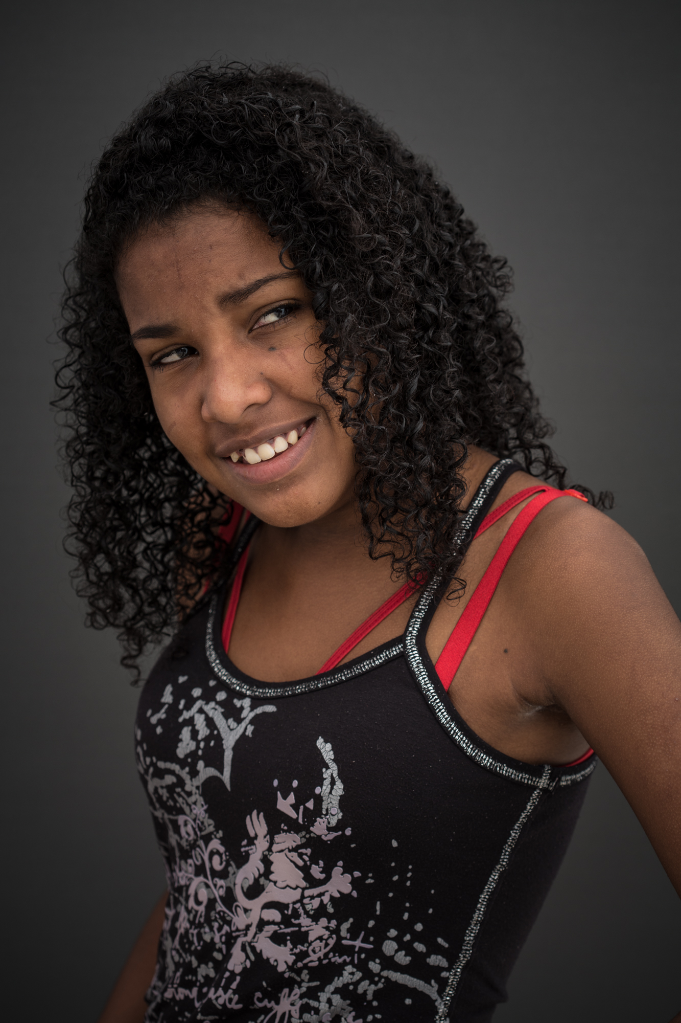 Adolescente negra sorri com olhar virado para a direita. Tem cabelos cacheados na altura dos ombros, Usa uma blusa preta com detalhes em branco e aparecem alças vermelhas da roupa de baixo.