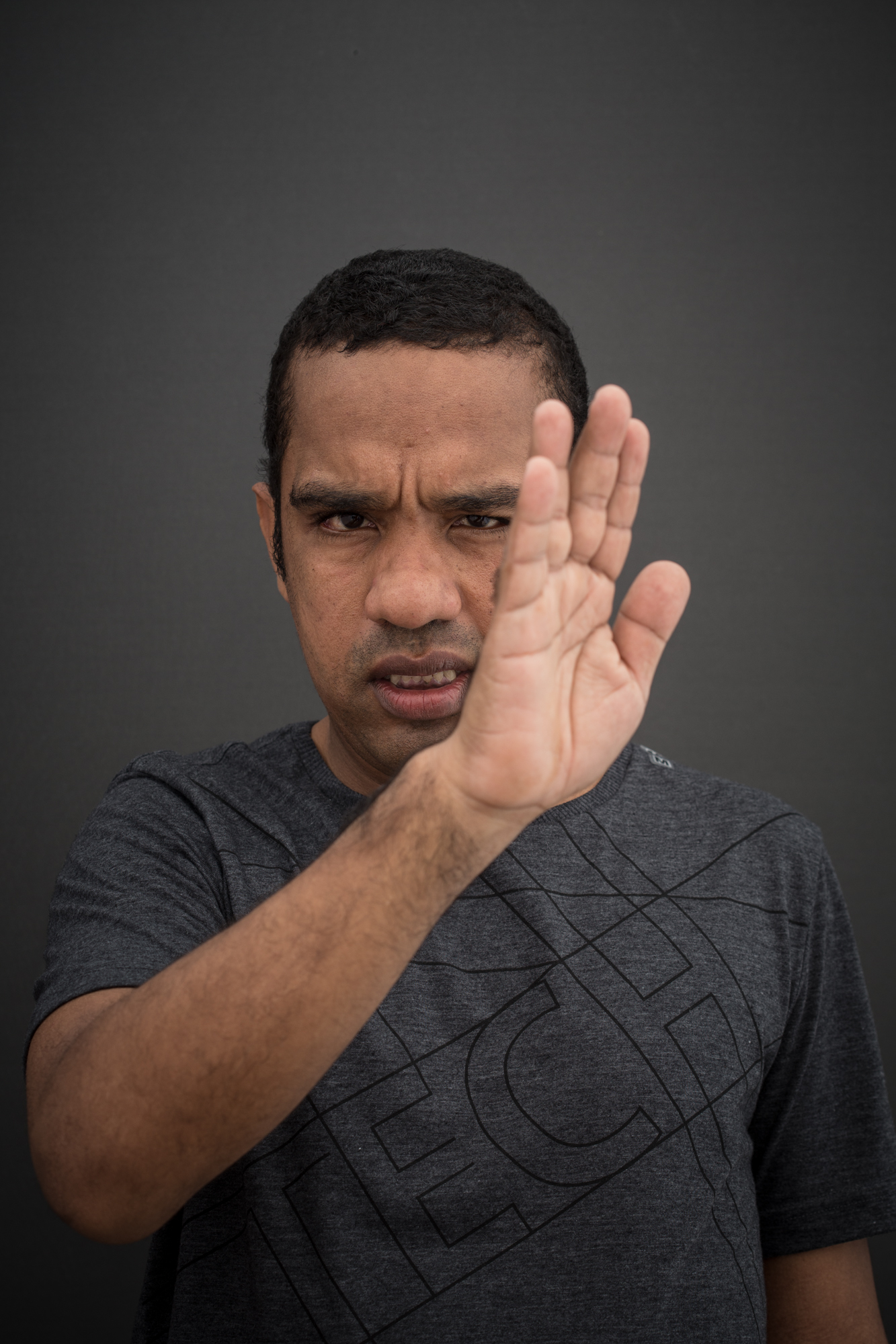 Homem negro olhando para a câmera com o rosto sério. Tem cabelo preto curto e usa uma camisa cinza escuro com linhas pretas. Ele está com a mão direita espalmada para o lado e erguida, cobrindo parte do seu rosto.