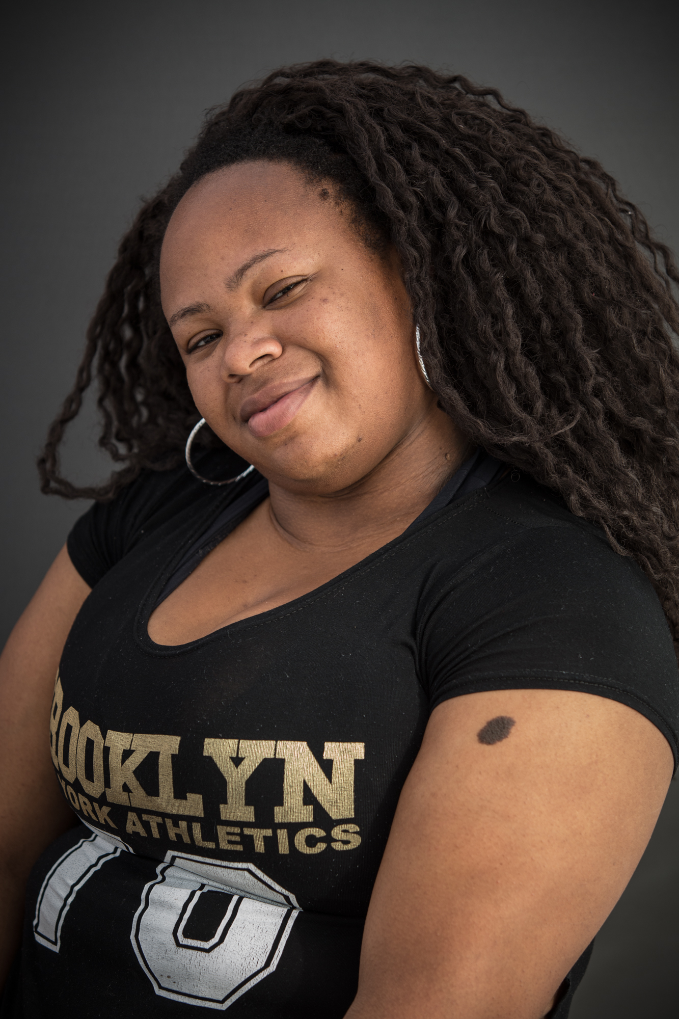 Jovem negra sorri em close. Usa cabelos com longas tranças e uma blusa preta com uma estampa onde se lê um pedaço do texto Brooklin Work Athletics em dourado e o número 70 em branco.