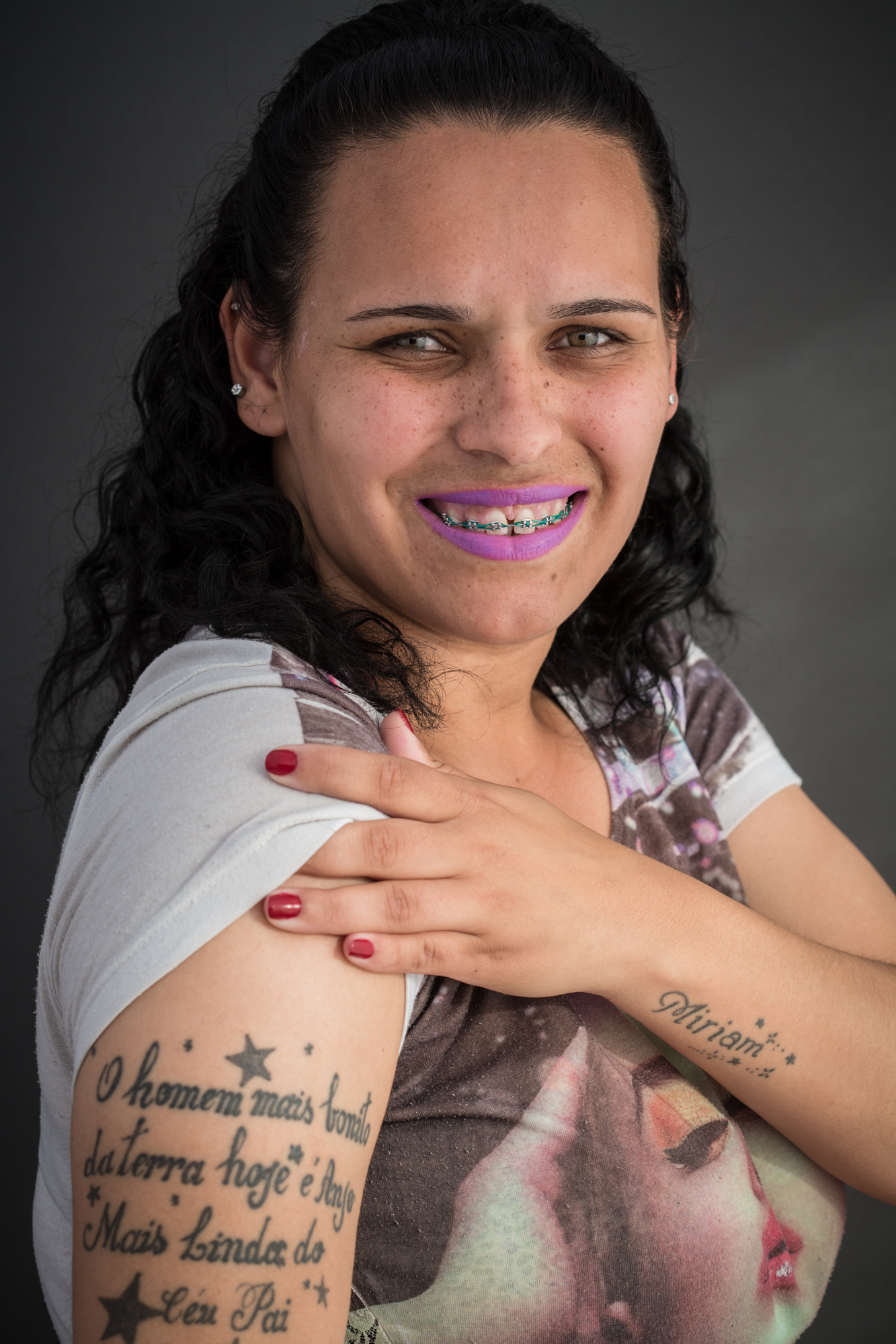 Mulher jovem sorrindo. Usa aparelho dentário, batom e camiseta branca com estampa de uma foto colorida. Com mão ela levanta uma das mangas e exibe uma tatuagem no braço com o texto: O homem mais bonito da terra hoje é anjo mais bonito do céu Pai.
