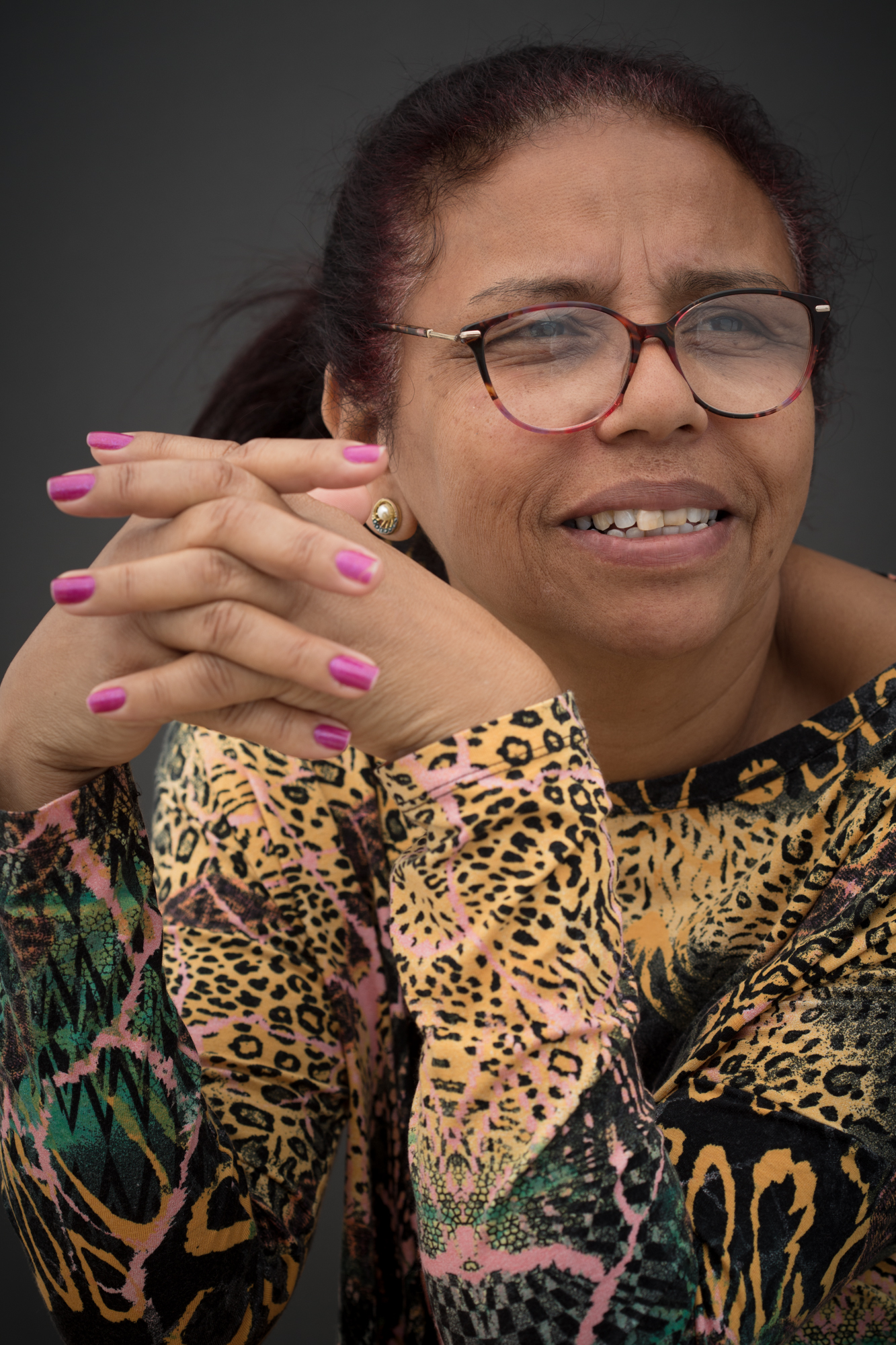 Close de mulher negra que sorri olhando para o lado. Usa óculos com armação em vermelho e preto, e uma blusa com estampa tigrada, com detalhes em verde e rosa. Está com as mão trançada a sua frente e tem as unhas pintadas de rosa.