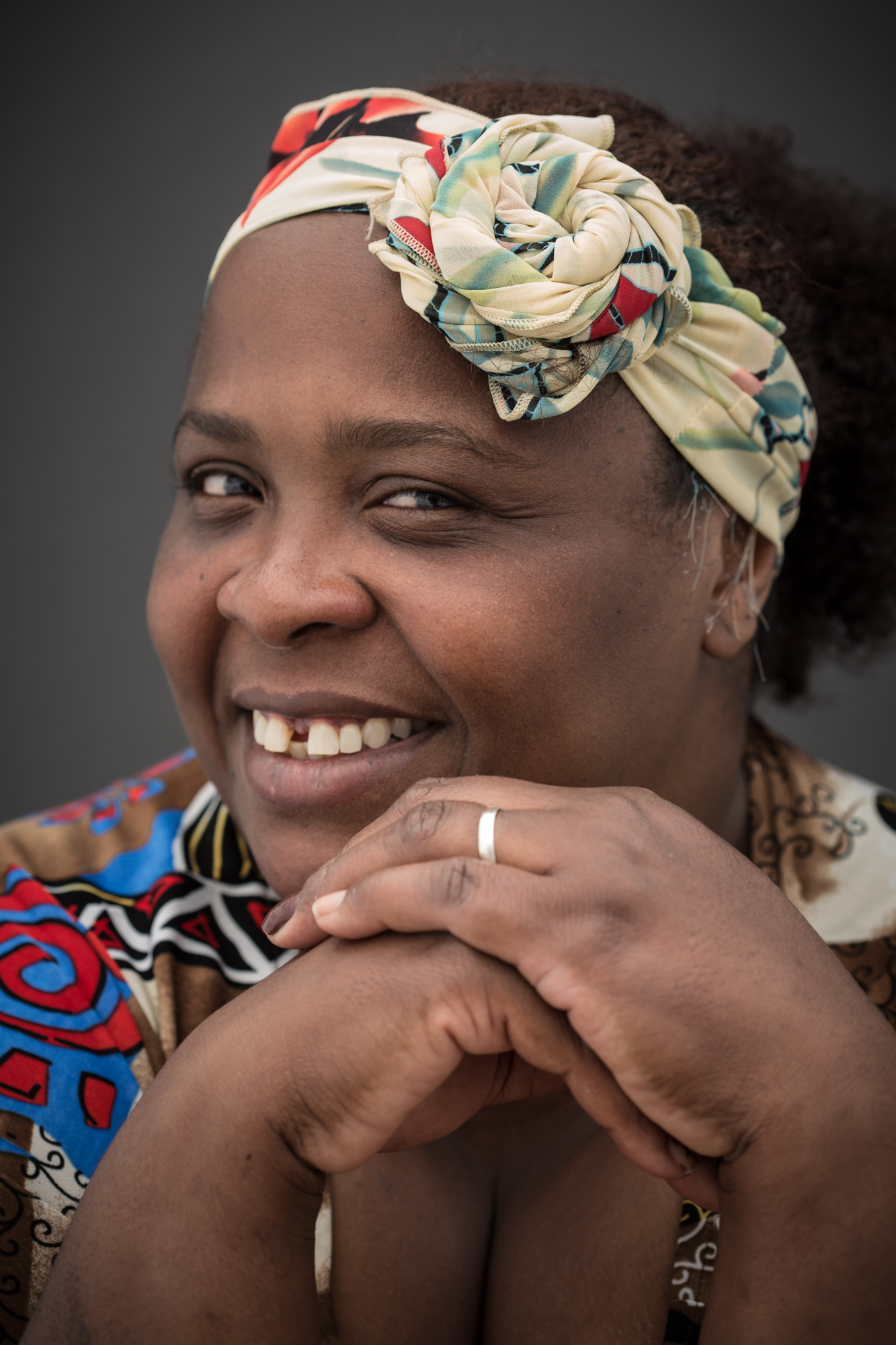 Close de mulher negra que sorri olhando para a câmera. Está com o rosto um pouco para o lado. Usa um lenço amarrado na cabeça com um arranjo em forma de rosa. Sua blusa tem estampas com padrões variados em vermelho, azul, preto, branco e dourado.
