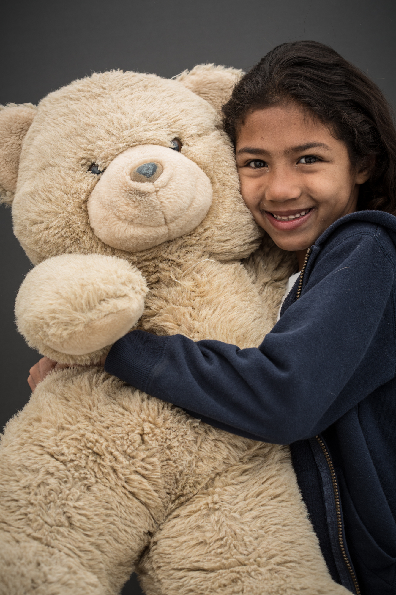 Criança parda sorri abraçando um grande urso de pelúcia bege.  Ela usa o cabelo comprido preto e veste uma jaqueta de algodão azul marinho com uma camiste branca por baixo.