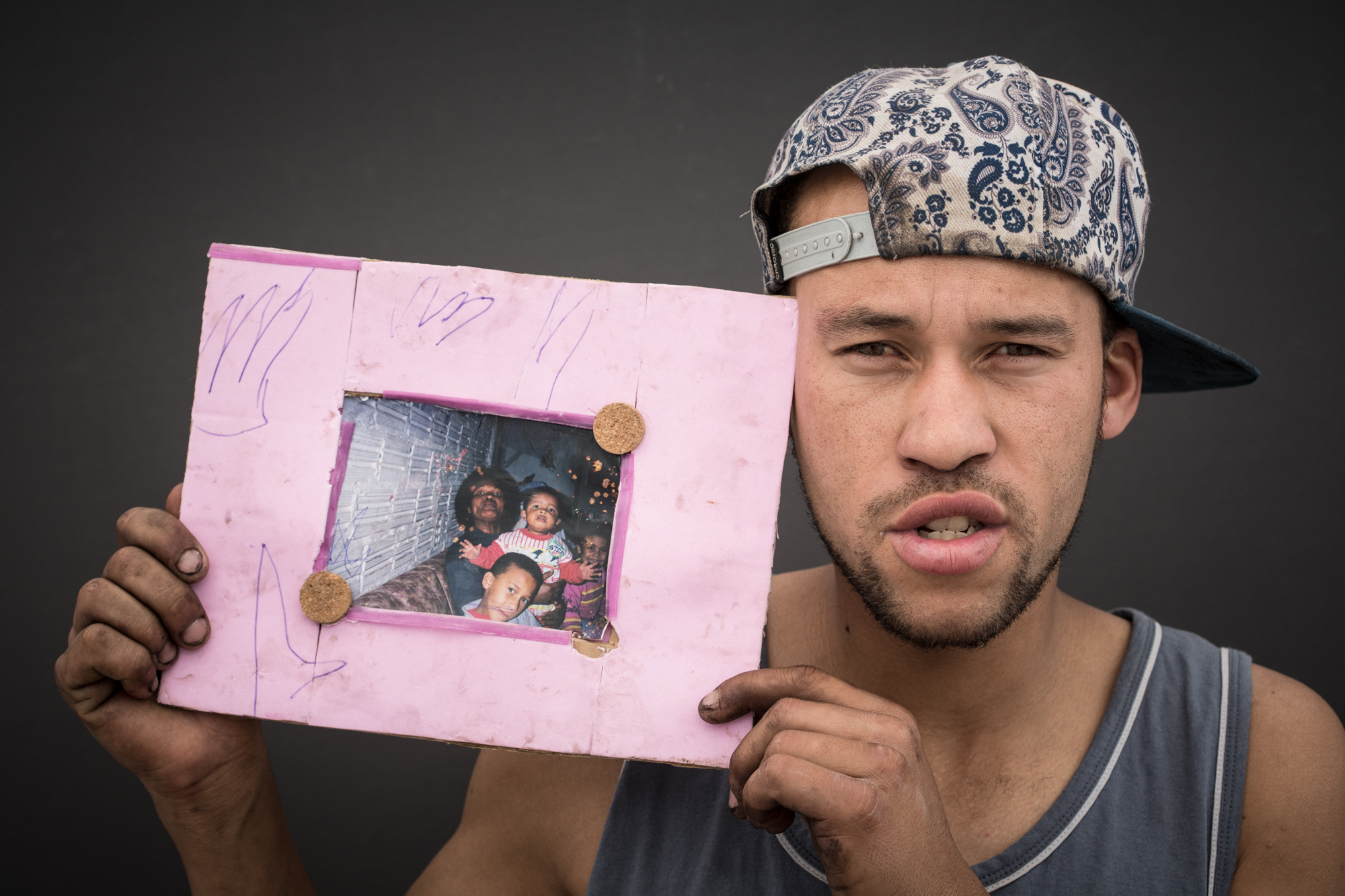 Jovem pardo segura um papel com uma foto colada no centro, onde ele aparece criança com sua mãe negra e dois irmãos menores. Ele usa um boné virado para o lado e uma camisa de física cinza.