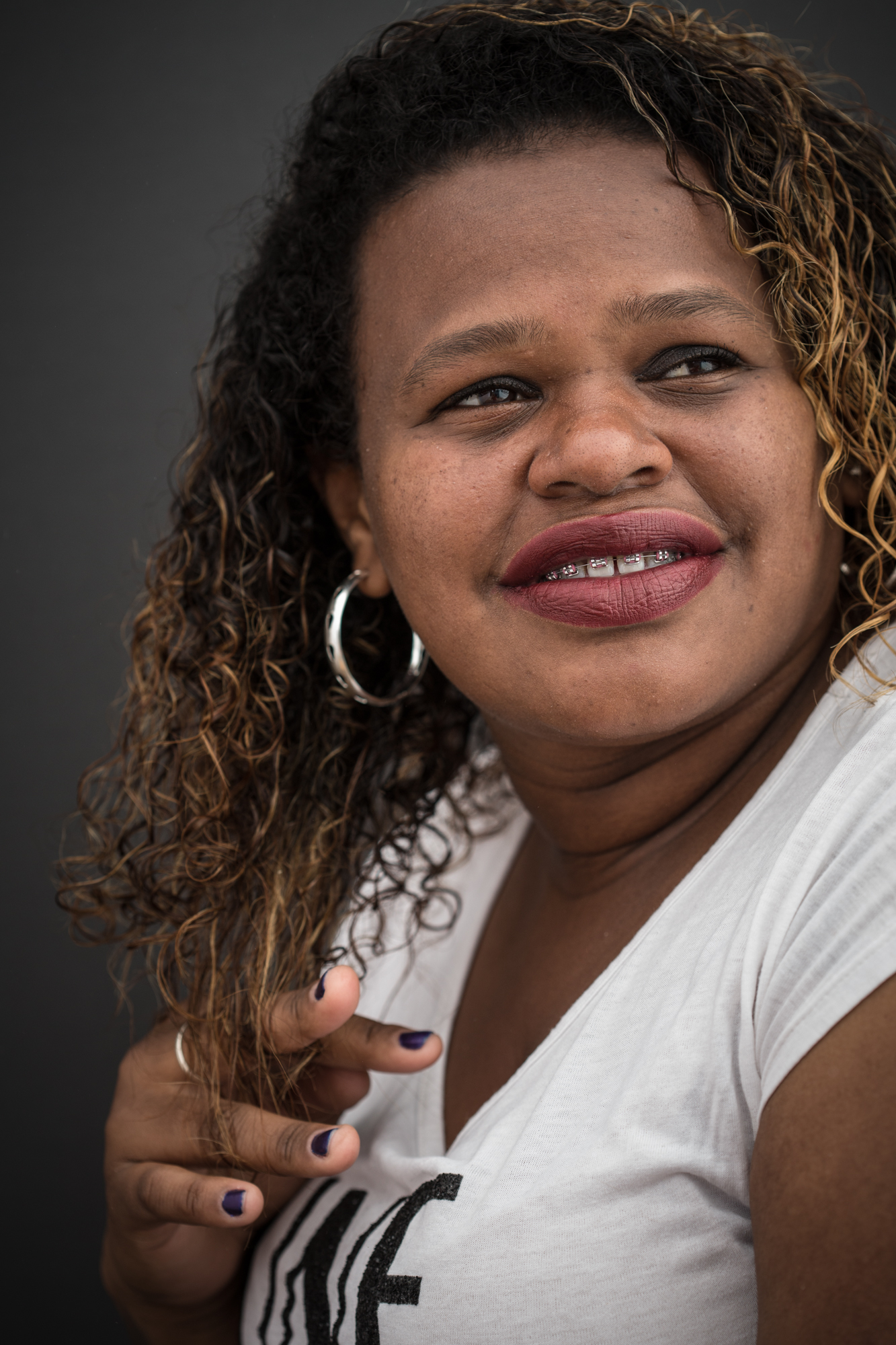 Close de mulher negra sorrindo olhando para o alto. Usa o cabelo preto cacheado longo com luzes douradas. os dedos da mão direita brincam com a ponta dos cachos. Usa uma blusa branca, brinco de argola e aparelho dentário.