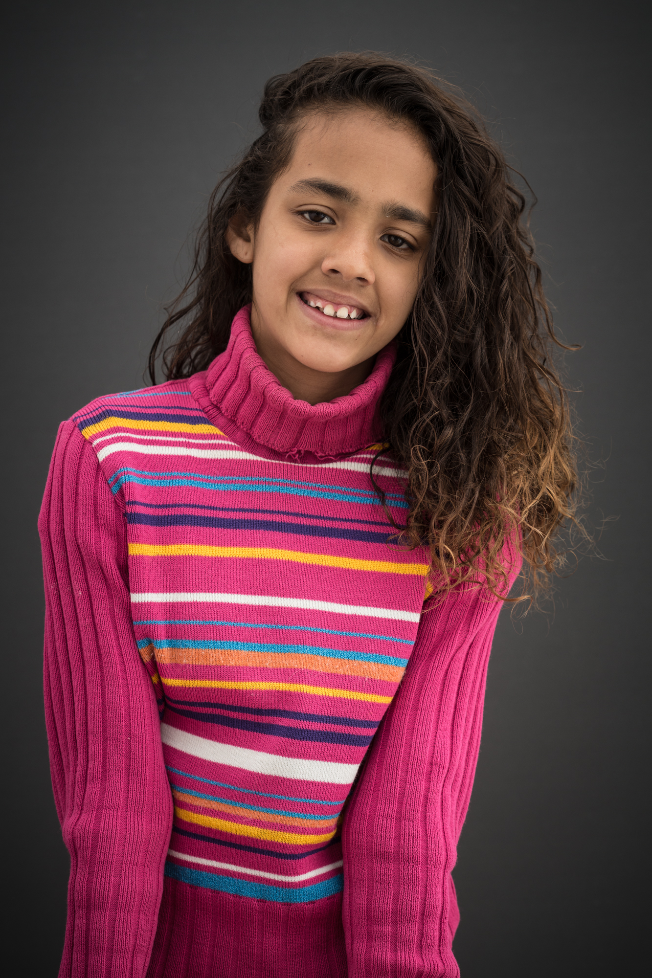Criança negra de cabelos longos e castanhos sorri. Usa um blusão rosa com listras em cores diversas.