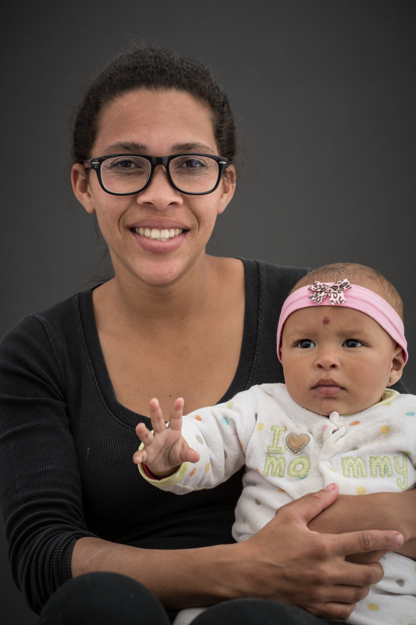 Mulher negra sentada segurando um bebê. Ela usa óculos e armação preta e blusa preta. O bebê tem um laço rosa na cabeça e usa uma blusa branca com desenhos coloridos.