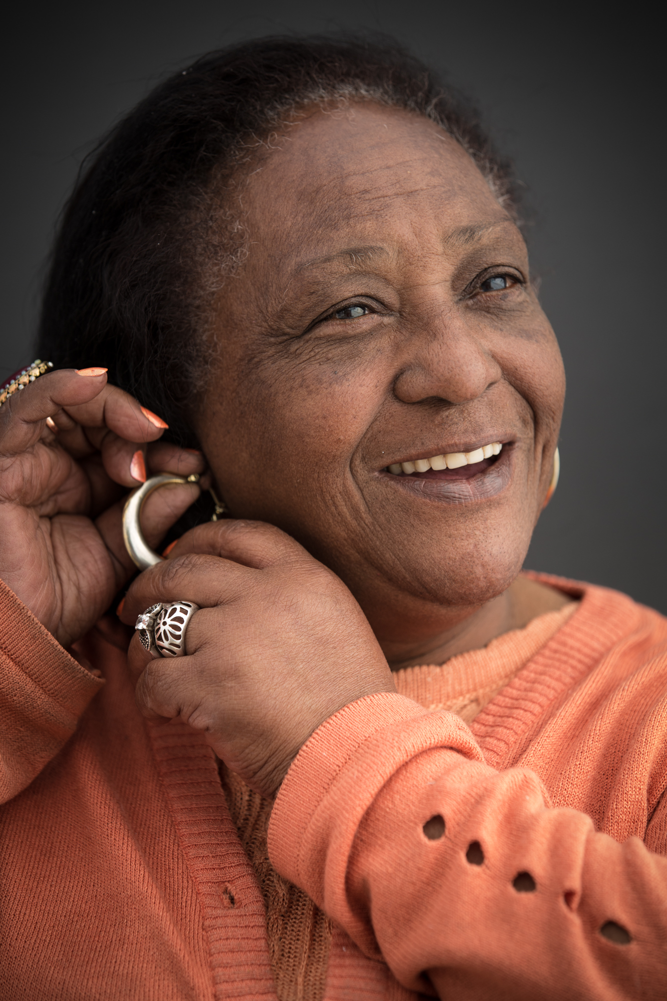 Mulher negra idosa ajeitando um brinco de argola dourado na orelha. Ela sorri. Usa um xale laranja, unhas pintadas de laranja e anéis dourados.