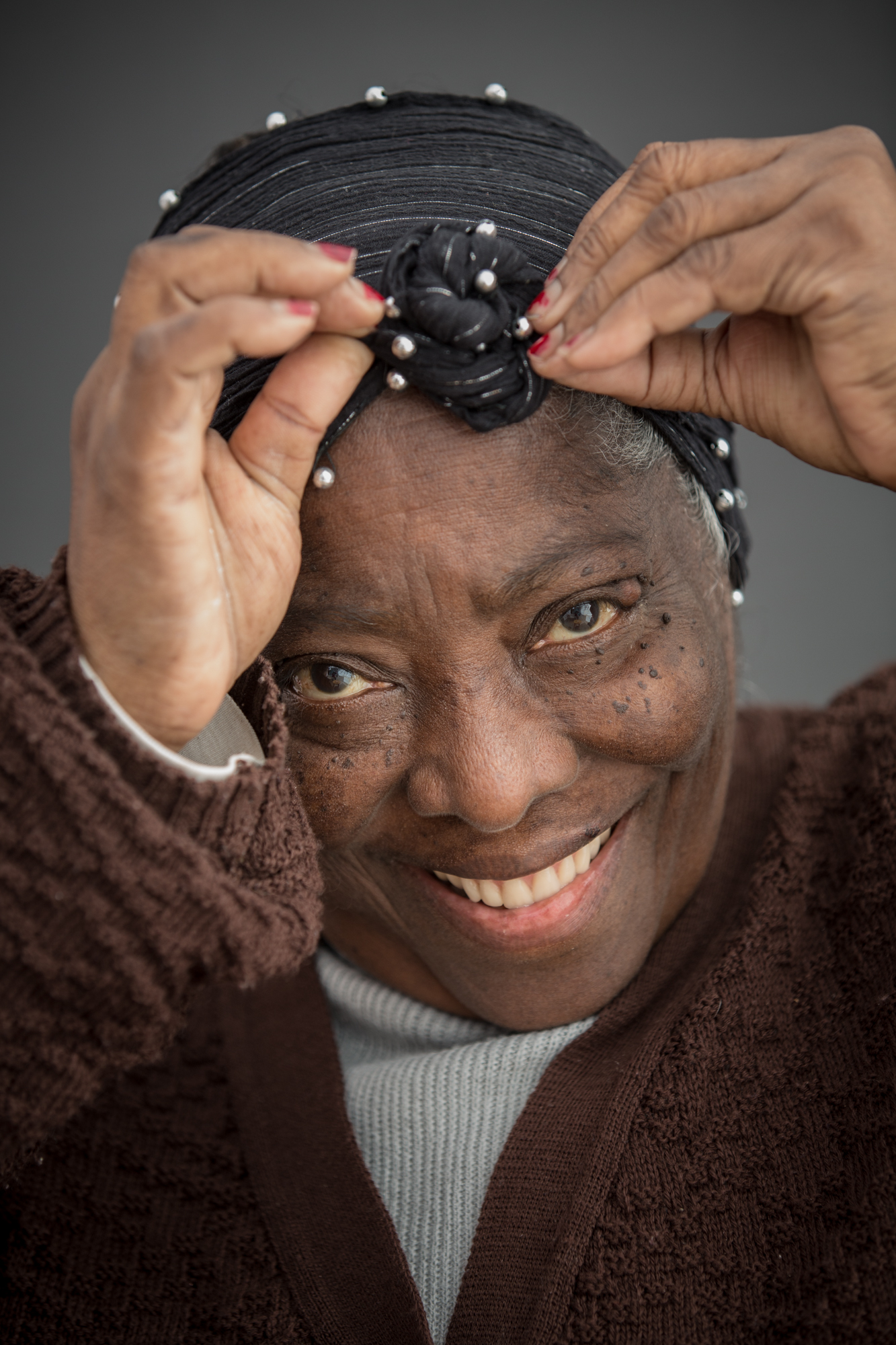 Mulher negra idosa ajeitando um lenço preto na cabeça. Sorri olhando para a foto. Usa um blusão branco sob um xale marrom.