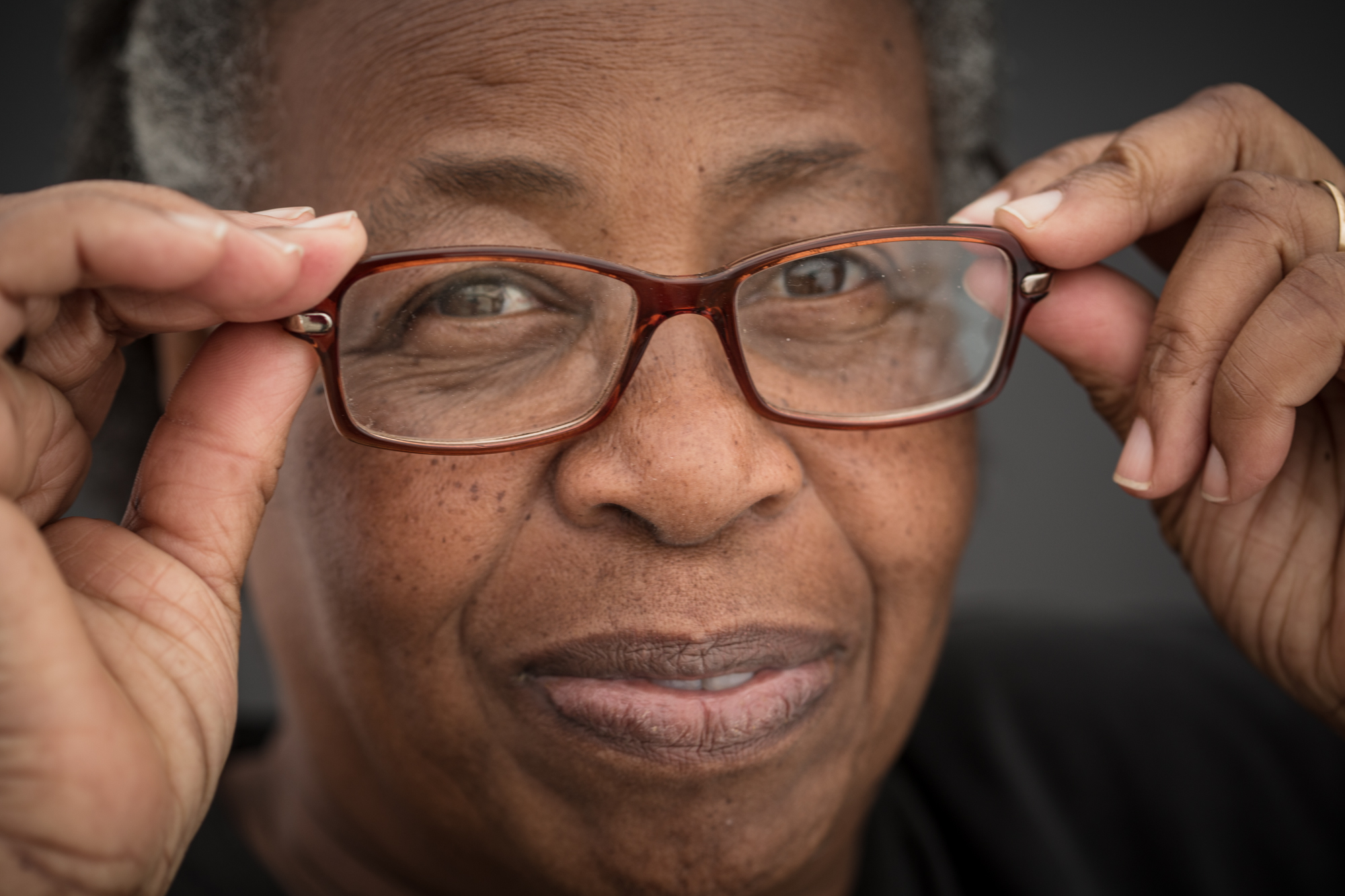 Close de mulher idosa negra ajustando os óculos no rosto. Ela tem o cabelo curto e grisalho. A armação dos óculos é vermelha. Ela sorri e segura os óculos com a ponta dos dedos.