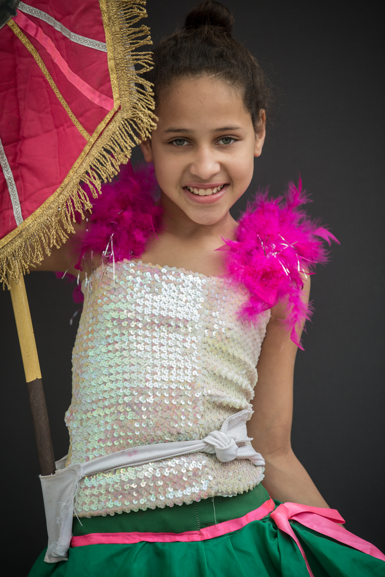 Menina segurando o estandarte da escola de samba. Aparece apenas o canto inferior do estandarte rosa na foto. A criança sorri para a câmera. Usa plumas rosas nos ombros, blusa branca com lantejoulas e saia verde com um laço rosa.