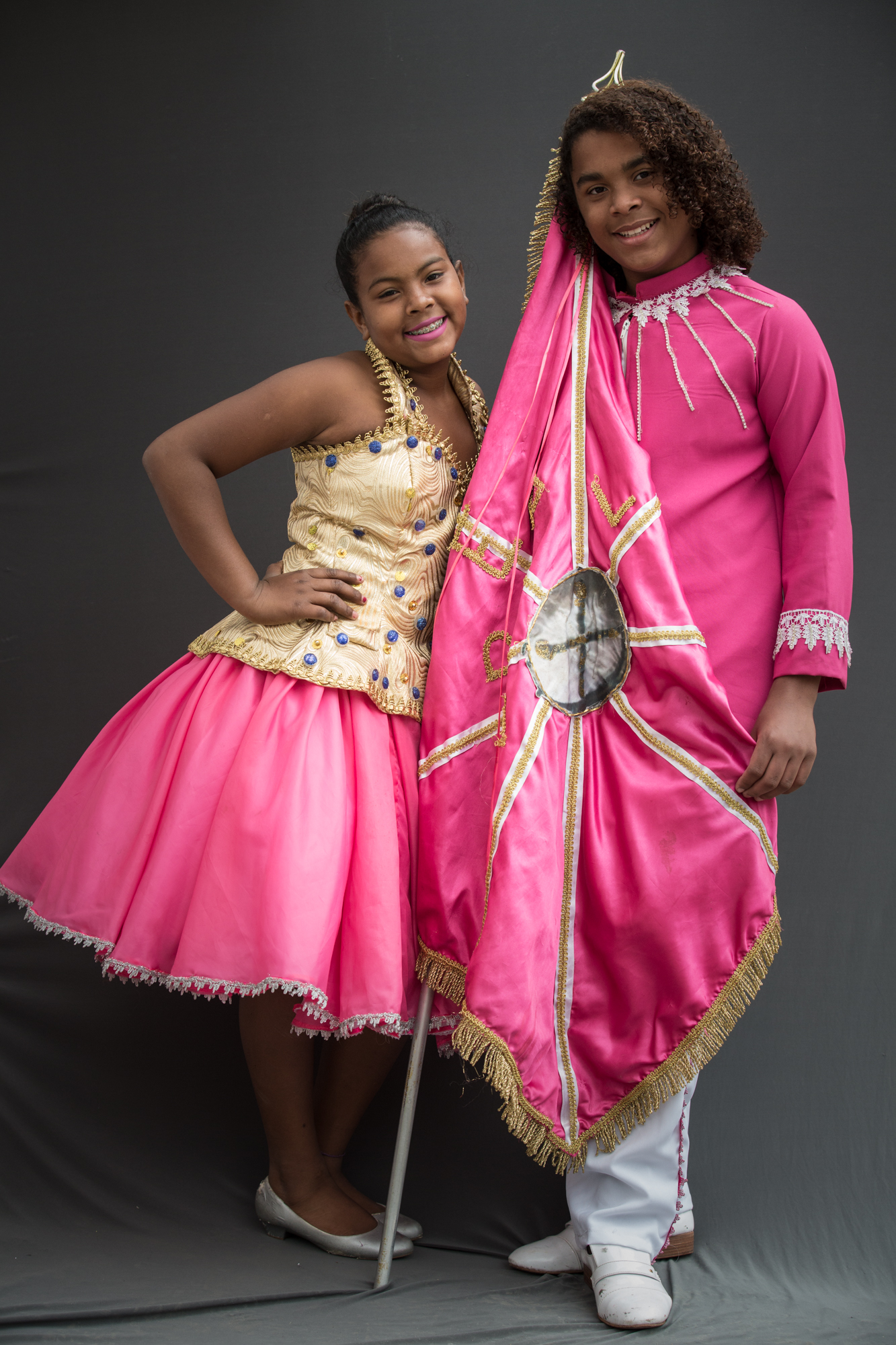 Casal de crianças com a bandeira rosa da escola de samba entre eles. A menina usa blusa dourada e saia rosa com sapatilha prateada. O menino usa calça e sapato branco e camisa rosa de manga comprida.