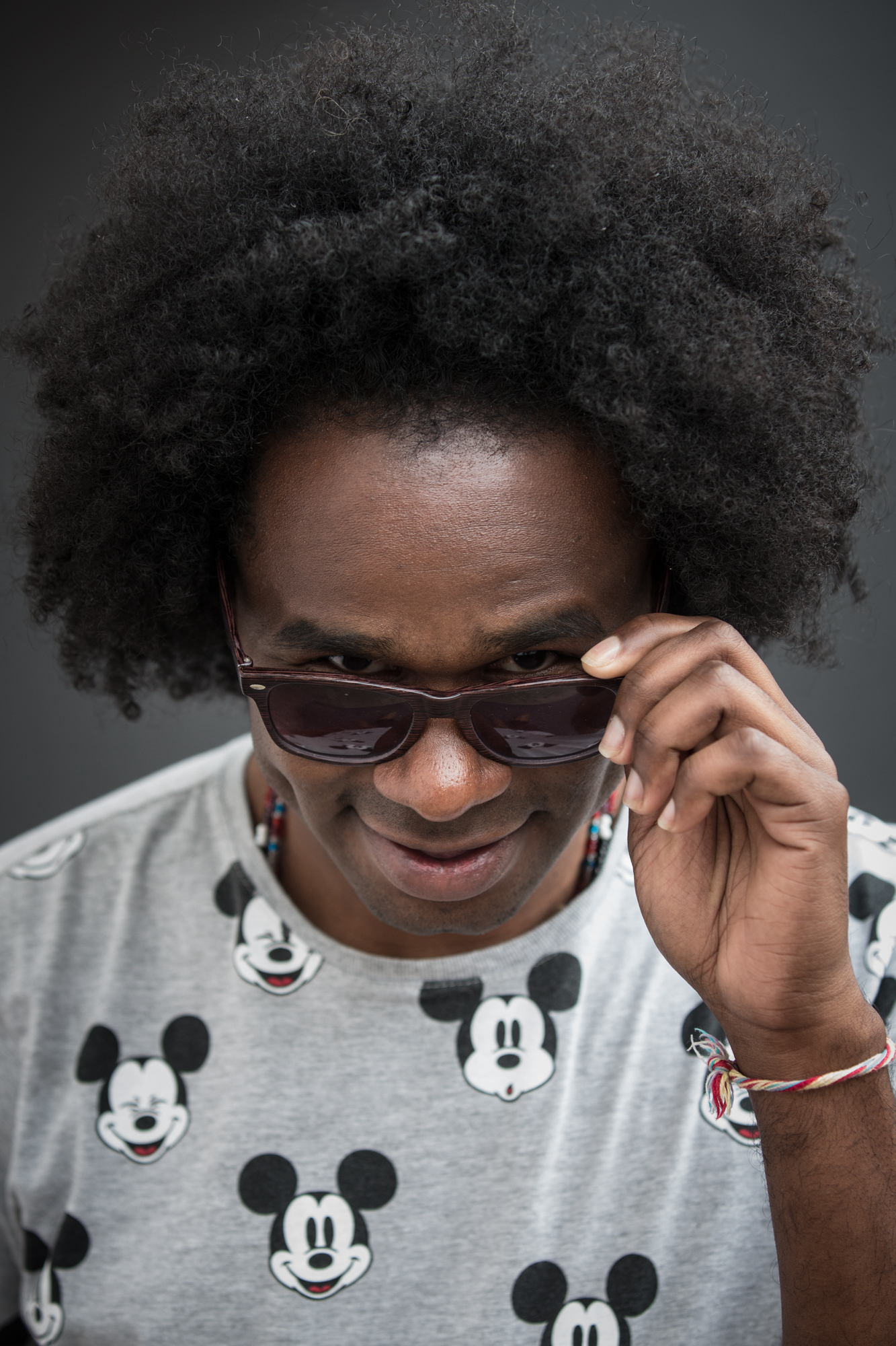 Close de homem negro com cabelo black power. Usa óculos escuros com armação marrom, segura eles com a mão esquerda e olha para a foto por cima das lentes, sorrindo. Usa camiseta cinza com estampas do Mickey e uma guia de santo amarrada no pulso.