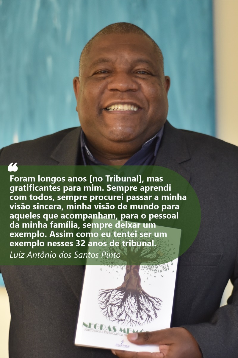Foto de Luiz Antônio Pinto sorrindo para a câmera e mostrando o livro Negras Memórias. Ele tem a pele e olhos negros. Seus cabelos são grisalhos e raspados curtos. Usa camisa escura e terno preto. Seu redor está desfocado.