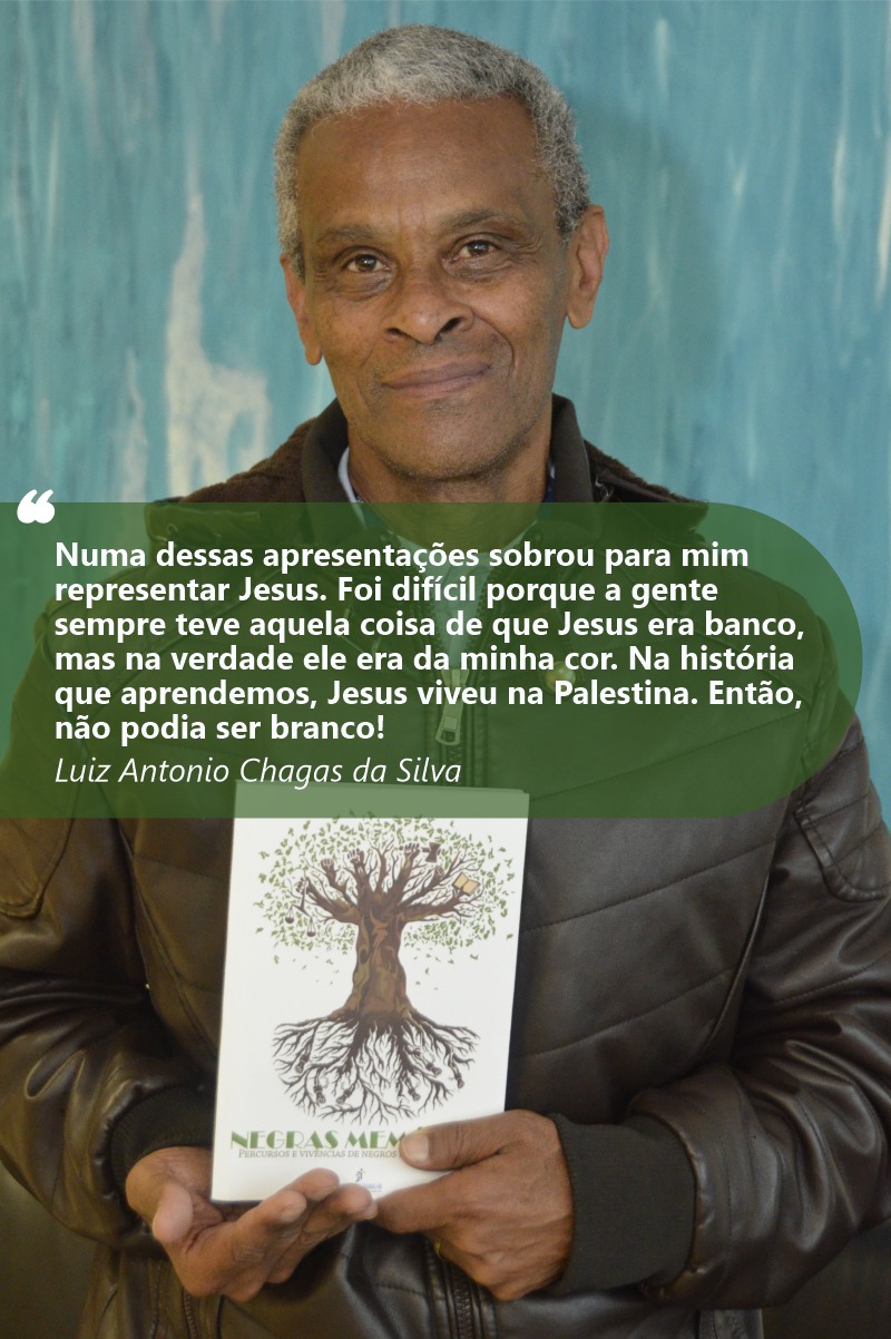 Foto de Luiz Antônio Chagas sorrindo para a câmera e mostrando o livro Negras Memórias. Ele tem a pele e olhos negros. Seus cabelos são grisalhos e curtos. Usa casaco preto. Seu redor está desfocado.