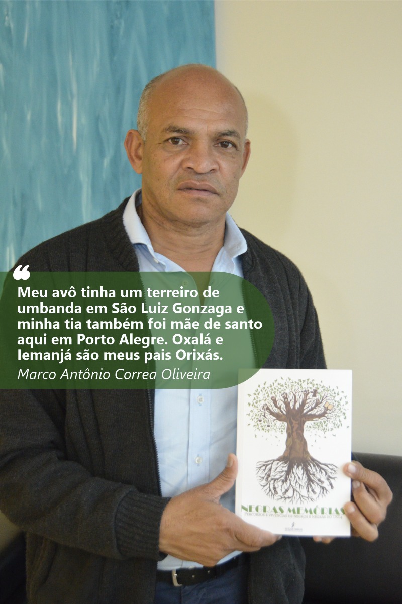Foto de Marco Antônio com olhando para a câmera e mostrando o livro Negras Memórias. Ele tem a pele e olhos negros. É calvo, com cabelo curtos e grisalhos. Usa camisa azul clara e casaco preto. Seu redor está desfocado.
