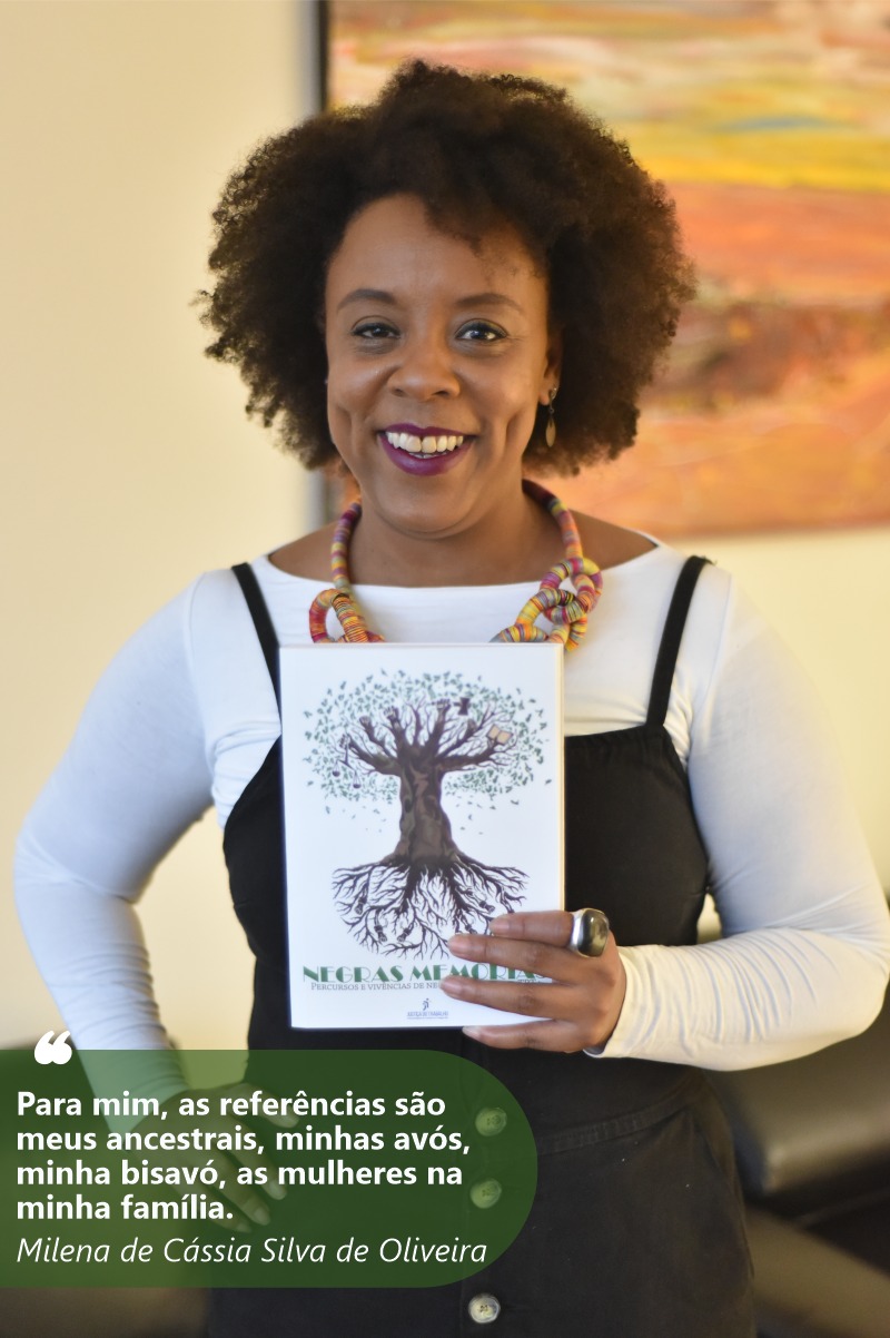 Foto de Milena sorrindo para a câmera e mostrando o livro Negras Memórias. Ela tem a pele e olhos negros.Seus cabelos são crespos e soltos, pouco longos. Usa colar colorido, blusa branca e macacão preto.