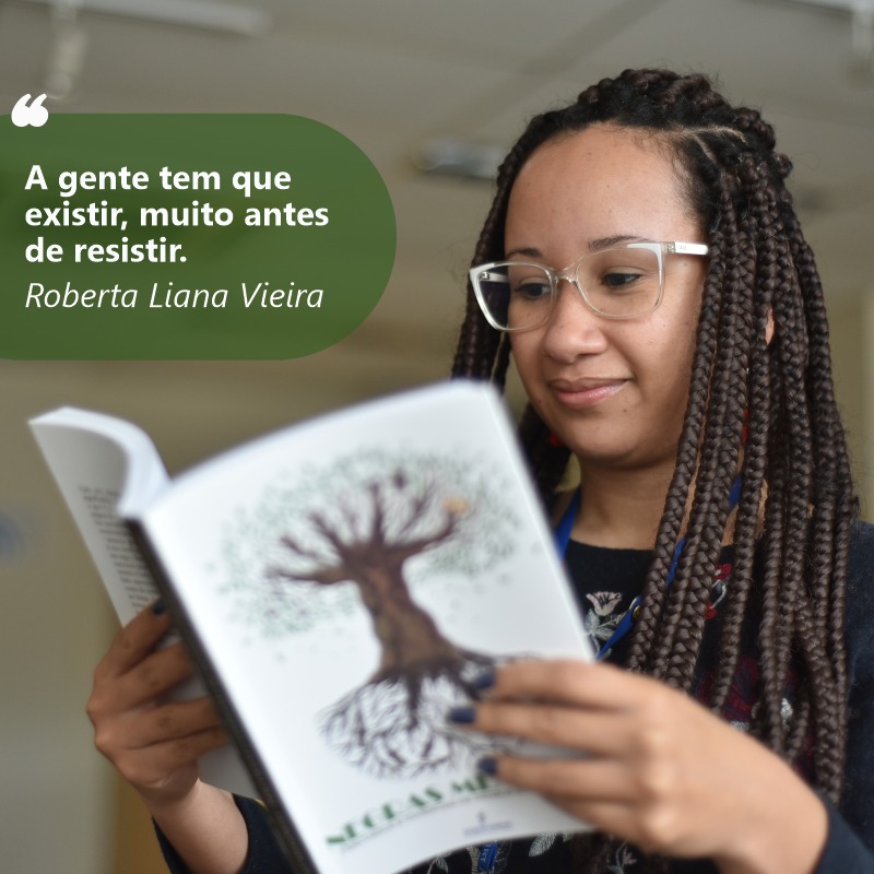 Foto de Roberta lendo o livro. Ela tem a pele e olhos negros. Tem os cabelos negros e presos em longas tranças aos lados do rosto. Usa óculos e uma blusa escura, com estampa floral. Tem as unhas pintadas de azul.