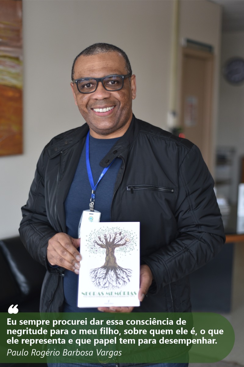 Foto de Rogério sorrindo para a câmera e mostrando o livro Negras Memórias. Ele tem a pele e olhos negros. Seus cabelos são negros e curtos. Usa camiseta azul e casaco preto. Seu redor está desfocado.