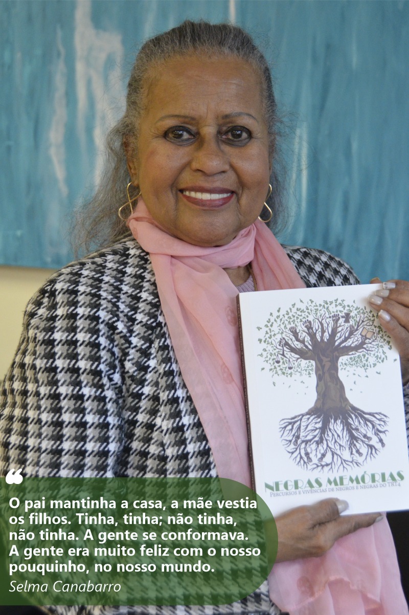 Foto de Selma sorrindo para a câmera e mostrando o livro Negras Memórias. Ela tem a pele e olhos negros. Usa um casaco xadrez e echarpe rosa. Seus cabelos grisalhos e lisos são longos e estão presos atrás da cabeça.