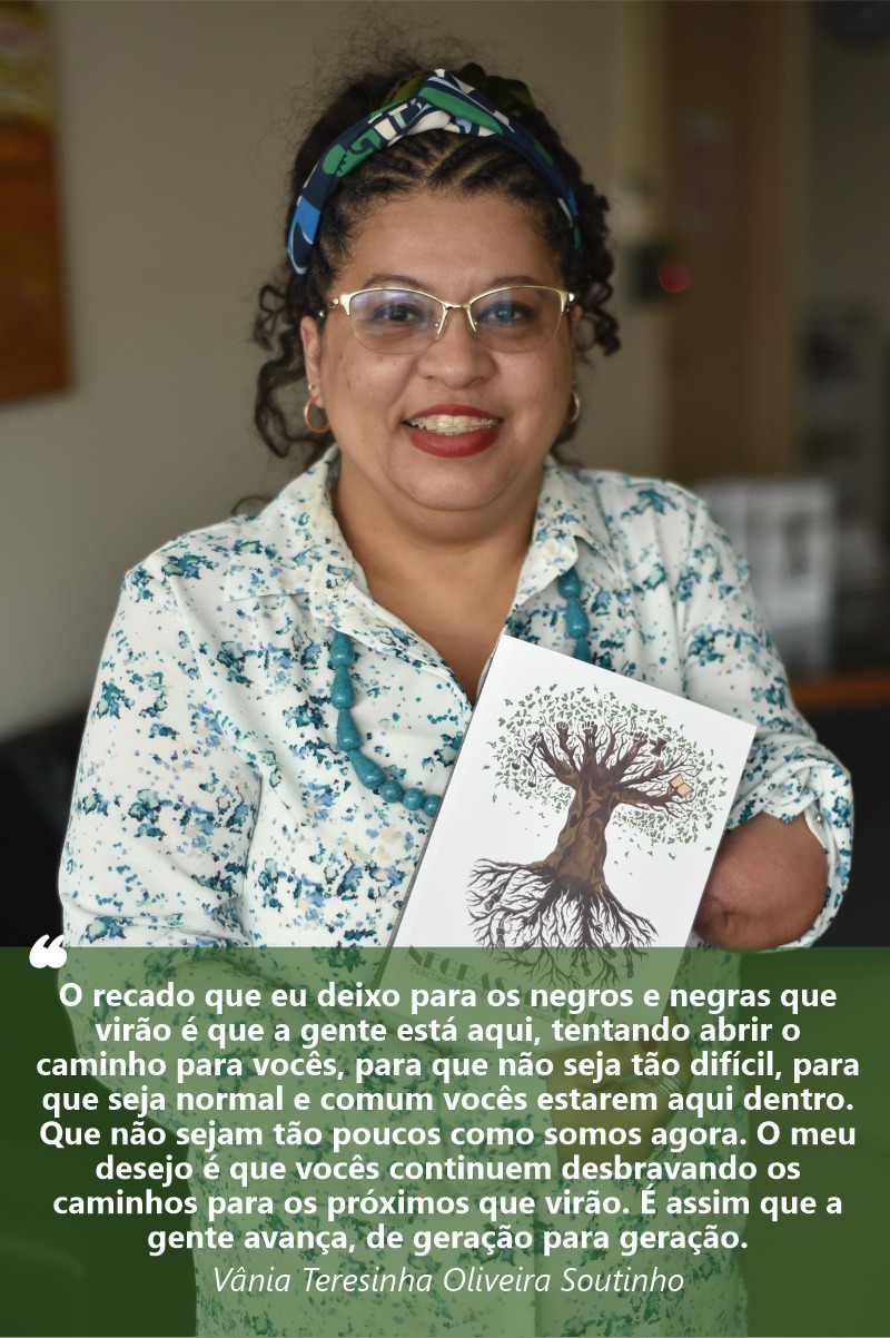 Foto de Vania sorrindo para a câmera e mostrando o livro Negras Memórias. Ela tem a pele e olhos negros. Seus cabelos longos e crespos estão trançados e presos por uma faixa colorida, no alto da cabeça. Usa uma camisa clara, com estampa de flores azuis.