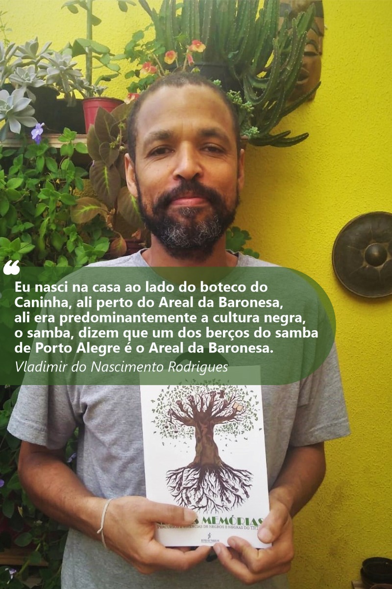 Foto de Vladimir sorrindo para a câmera e mostrando o livro Negras Memórias. Ele tem a pele e olhos negros. Sua barba e cabelos são negros. Usa camiseta cinza. Ao seu redor, aparecem plantas e um muro amarelo.