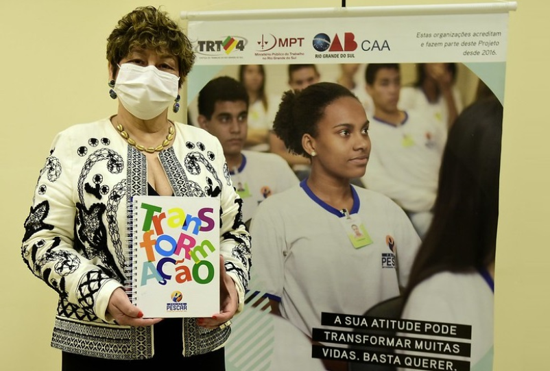 Foto posada da presidente do TRT-RS Carmen Gonzalez. Ela segura e mostra para a câmera um caderno do Pescar, com a palavra "transformação" escrita na capa, em letras grandes e coloridas. Ao lado dela, um banner do projeto.