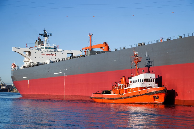 Foto ilustrativa de um navio de grande porte no oceano. Junto dele há o barco rebocador, uma embarcação de porte pequeno.