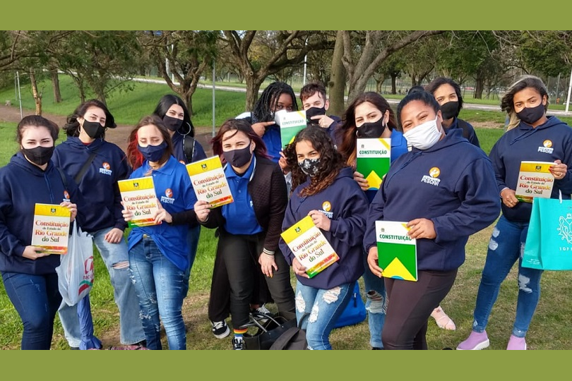 Fotos posada dos jovens da turma do TRT. Eles usam máscaras e o uniforme (casaco e/ou camiseta polo azuis). Estão sobre a grama de um parque, com árvores à volta. Exibem para a câmera exemplares das constituições federal e estadual.