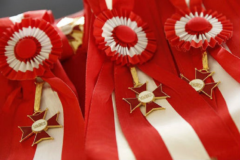 Foto ilustrativa do distintivo da Comenda do Mérito Judiciário. São pequenas medalhas vermelhas em formato de cruz, companhadas de uma faixa de tecido nas cores vermelha e branca.