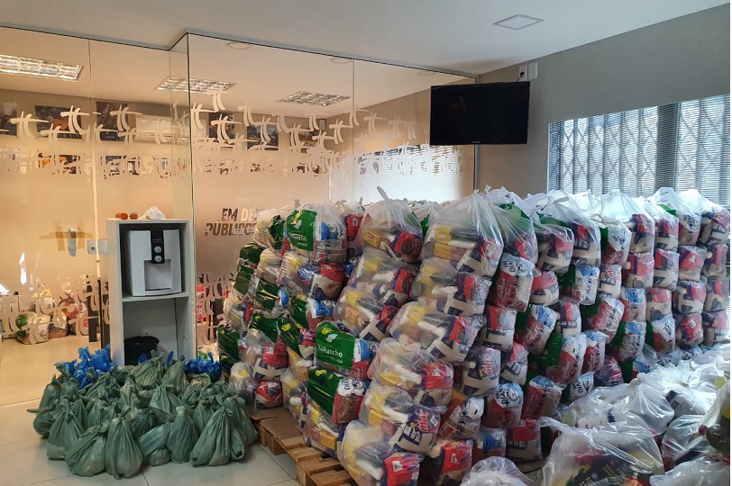 Foto mostra cestas básicas e kits de higiene organizados para a distribuição. A sala é clara e as sacolas estão enfileiradas e empilhadas.