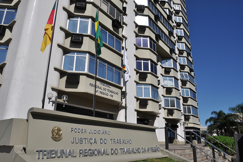 Fachada do Prédio-Sede do TRT-RS. A foto mostra em primeiro plano um letreiro com o nome do Tribunal e as bandeiras do Brasil, do RS e de Porto Alegre. Ao fundo aparece a fachada do prédio e o céu azul sem nuvens.