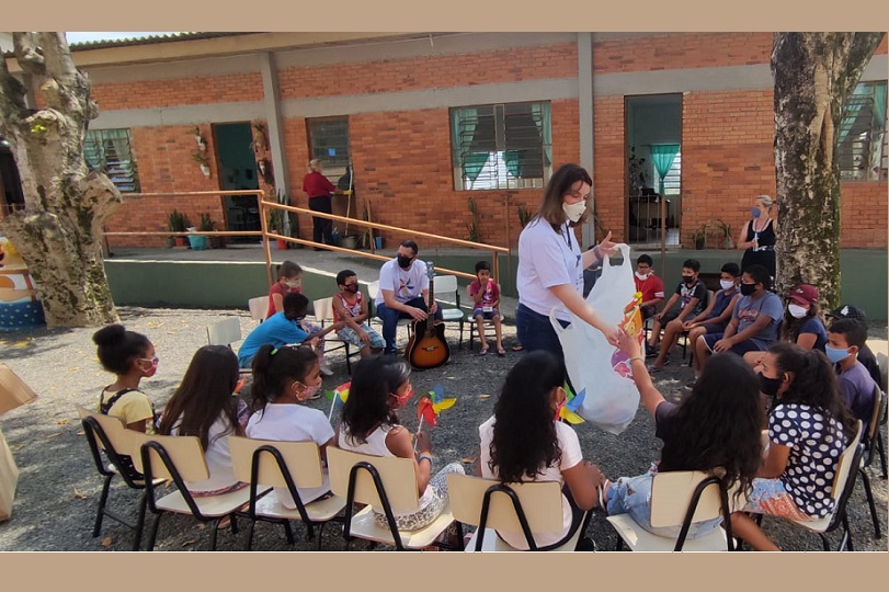 A servidora Angie Miron está ao centro de uma roda de crianças. Ela distribui os cataventos coloridos, símbolo do combate ao Trabalho Infantil.  Daniel Germano, compositor da música "Lugar de Criança é na Infância, está sentado e segura um violão.