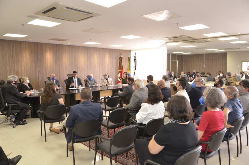 Foto em plano aberto da reunião. Mostra a mesa oficial e demais participantes sentados em cadeiras colocadas em frente à mesa.
